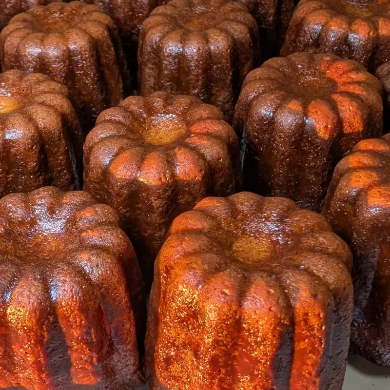 Rows of fluted, deep-brown cannelés with glossy, caramelized ridges, standing on a light tray