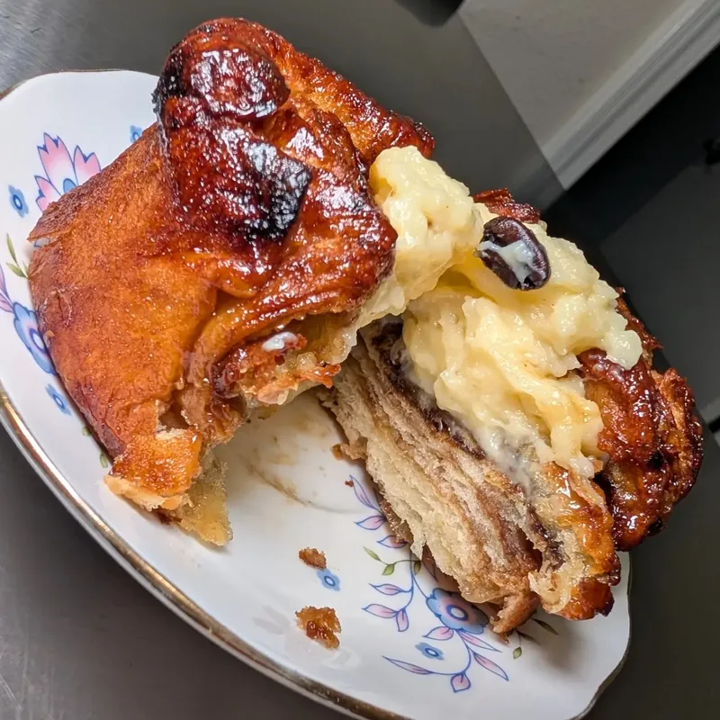 A glossy, dark-caramelized pastry broken open on a floral porcelain plate, showing flaky layers and a pale yellow cream filling with a dark bean on top