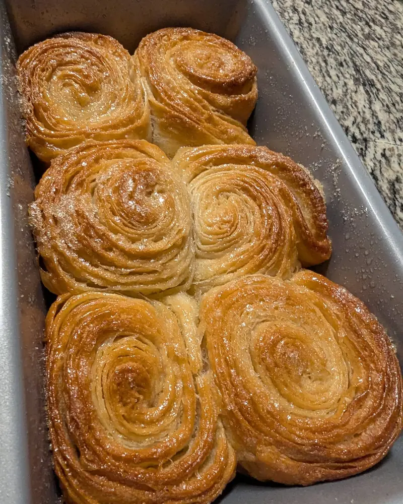 Six golden, spiral-layered pastries packed into a metal baking pan, dusted with sparkling sugar, on a speckled granite counter