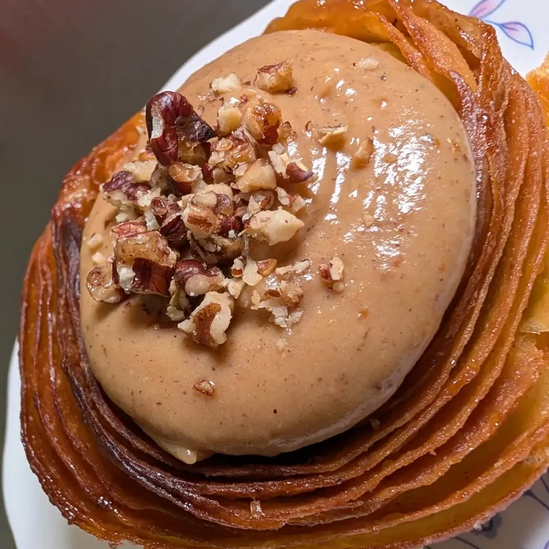 Close-up of a flaky, caramelized pastry topped with a smooth dome of light-brown cream and chopped pecans, on a floral porcelain plate